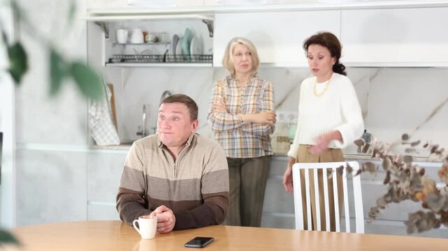 Upset middle-aged man sitting at table while two female family members scolding him standing behind in the kitchen