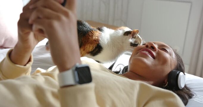 African American woman holding phone on bed as calico cat entering nuzzling face seeking affection