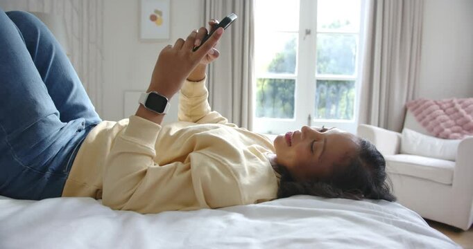 Adult African American woman lying on bed holding phone, tapping, reading texts, smartwatch visible