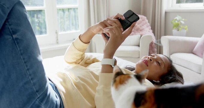 African American woman in bed using phone while calico cat climbing onto chest causing adjustments