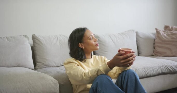 African American woman sitting floor by grey sofa in cream top, jeans holding mug close smiling