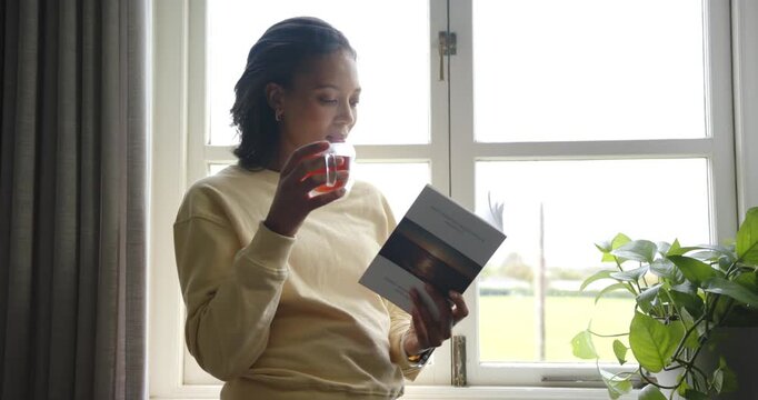Adult woman in yellow top holding book by window resting, reading, turning pages and sipping drink