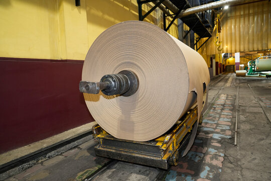 Massive industrial paper roll on a yellow wheeled trolley sits on a concrete floor, ready for transport within a paper mill production area filled with machinery