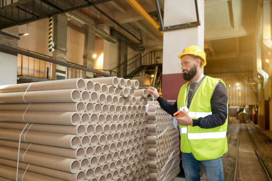 Man in protective gear diligently checks a large quantity of tightly packed cardboard tubes, managing inventory with his phone in a production area