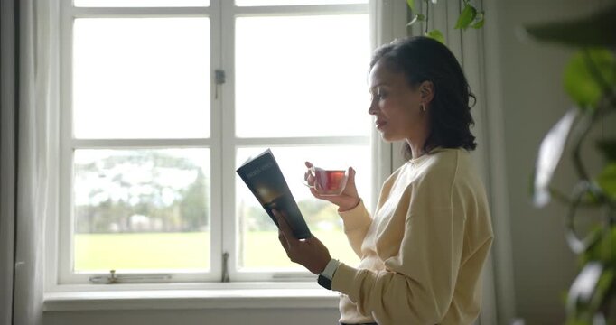 African woman standing by home paned window holding booklet teacup gazing at sun, copy space