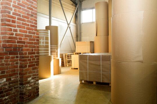 Large rolls of brown paper and stacks of cardboard boxes sit on pallets inside a warehouse, illuminated by natural light entering through a window