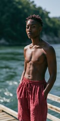 Fototapeta premium Shirtless young man standing on a wooden dock by a river wearing red shorts for portrait and lifestyle background