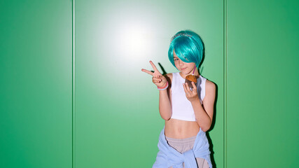 Young girl with vibrant blue wig and tiara holding a cupcake and making peace sign in front of green © khmelev