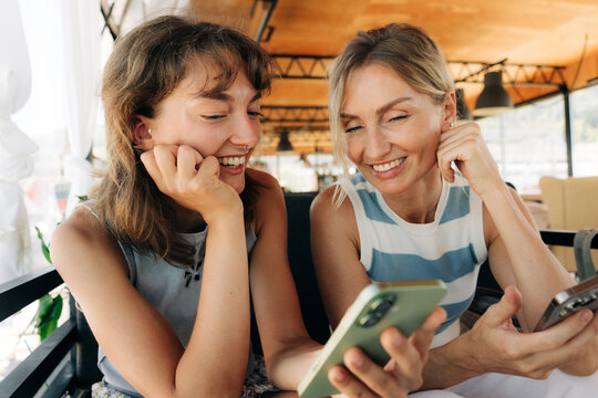 Two women of different ages are sitting in a caf&eacute;, scrolling through a social media feed on a phone.