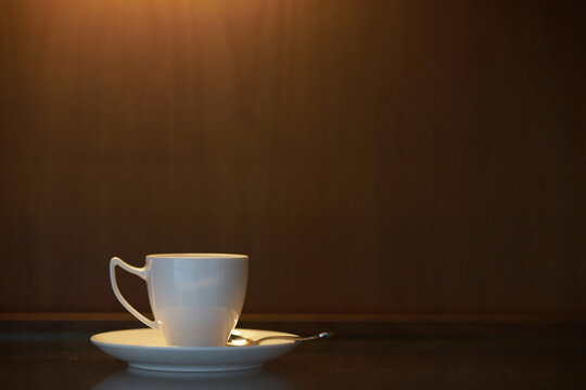 A white ceramic coffee cup and saucer sitting on a dark wooden table surface with warm atmospheric lighting casting down on it.