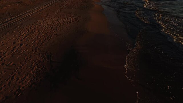 Dark ocean waves meeting textured sandy shoreline under warm sunset light with dramatic coastal contrast