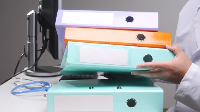 Doctor hands organizing a stack of colorful ring binders on a desk, representing healthcare administration, patient information management, and diligent record keeping in a clinic office