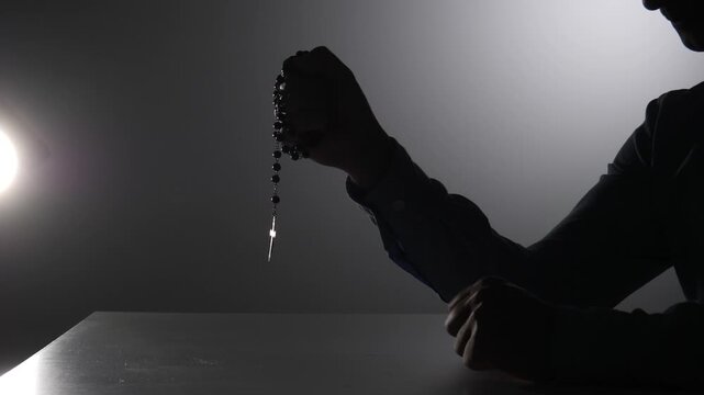 Person's silhouette holding a rosary with a cross in their hand, praying in a dark environment with a bright light on the side, representing faith and spirituality