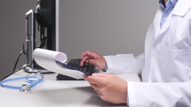 Physician wearing a white coat and holding a medical chart while typing patient information on a computer, indicating modern healthcare administration and digital record keeping