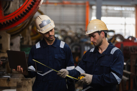 Serious industrial worker and engineer man wearing hardhat check heavy equipment with precision measuring tool and laptop computer inside manufacturing factory