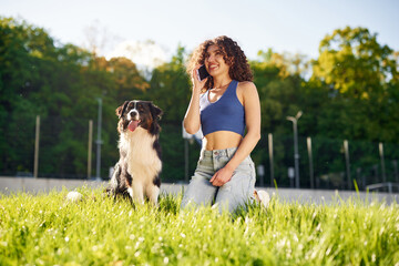 Fototapeta premium Young woman with curly hair kneeling on grass while talking on smartphone beside a black and white dog in a sunny park setting with trees in the background