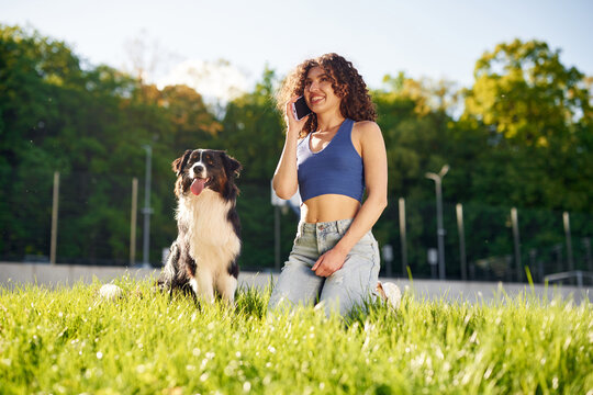 Young woman with curly hair kneeling on grass while talking on smartphone beside a black and white dog in a sunny park setting with trees in the background