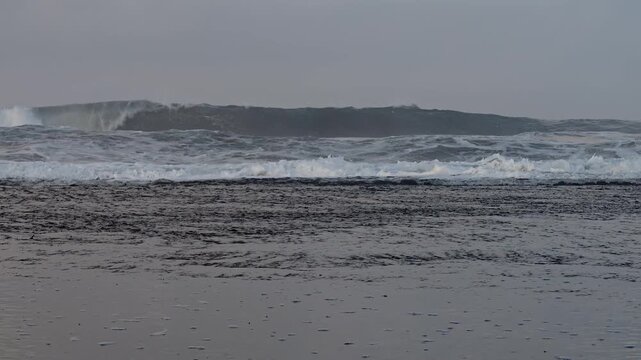 Big ocean waves rolling and crashing with white foam on dark wet rocky shore under cloudy sky, powerful sea water splashing in slow motion, scenic coastal nature background with natural ambient sound