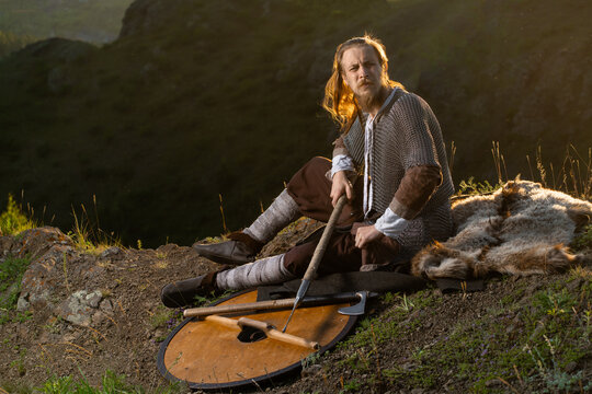 A rugged Viking warrior rests on a hillside with his axe and pelt, gazing thoughtfully into the distance.