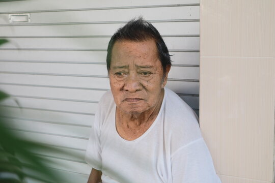 Senior man stands calmly against shuttered wall. His gaze holds quiet solemnity, unbothered. White t-shirt contrasts with subtle background tones. Natural light casts soft shadows across his face