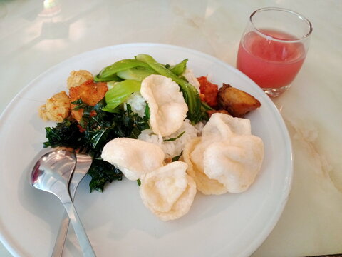 Indonesian mixed rice dish (Nasi Rames) served with crackers, green vegetables, side dishes, and a glass of fresh guava juice on a white plate.