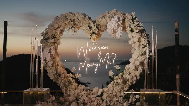 Romantic "Will you Marry Me?" neon sign glowing at twilight inside a large white floral heart arch. This exquisite coastal proposal setup features elegant candlelight and stunning ocean views.