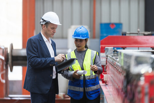 Manager and female worker engineer holding tablet during serious discussion about manufacturing inspection inside large industrial factory with heavy equipment