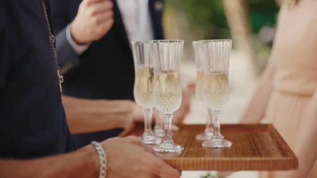 A waiter carrying a wooden tray with elegant crystal champagne flutes filled with sparkling wine, serving guests at a luxury outdoor wedding reception or upscale corporate party.