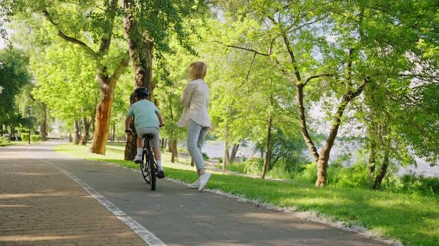 Mother claps as young boy rides bicycle on sunny park path