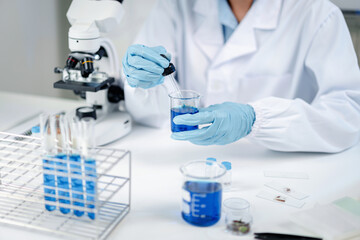 Fototapeta premium Scientist in white lab coat and blue gloves using dropper pipette to add solution into beaker of blue liquid, with microscope, test tubes and specimen slides on laboratory table