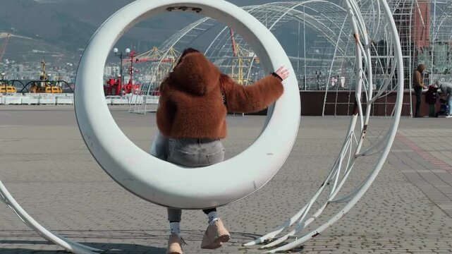 Anonymous female in faux fur coat swinging on large white ring structure at coastal town plaza overlooking industrial harbor cranes and mountain range on clear sunny afternoon casual lifestyle shot