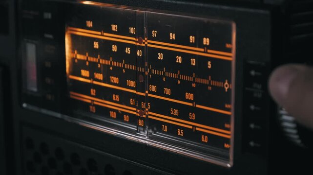 A hand turns the tuning knob on a vintage radio, moving the needle across the backlit analog dial. Concept of nostalgia, searching for information, retro technology, and communication.