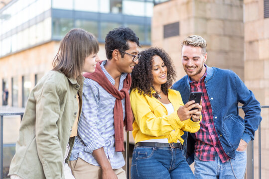Happy multiethnic friends laugh while sharing smartphone content in a modern city setting. Young millennials and gen z gather close outdoors. Digital friendship concept