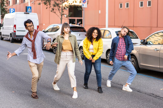 Goofy diverse friends making funny dance moves on a city street. Multiethnic millennials and gen z act silly together near parked cars. Humor concept