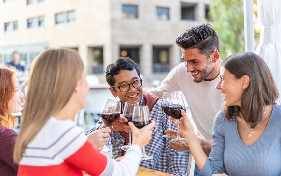 Diverse friends clinking glasses of red wine during a cheerful outdoor aperitif. Multiethnic millennials and gen z welcome a friend to the table. Urban hospitality concept