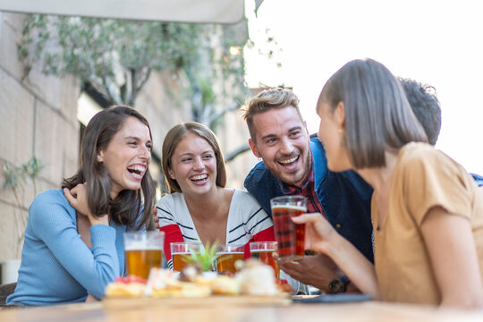 Young friends laughing over beers and appetizers at an outdoor city cafe. Millennials and gen z enjoy a relaxed terrace conversation. Happy hour concept