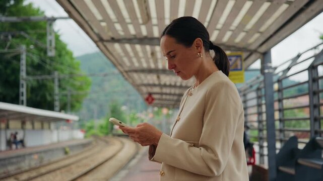 An adult woman uses her mobile phone while standing on a train station platform, waiting for public transport.