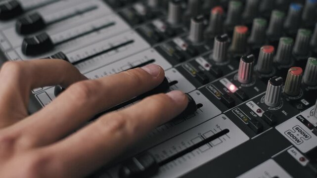 High angle close-up of a sound engineer's hand pushing two channel faders on a professional mixing board. The shot features detailed decibel markings and multiple sliders in shallow focus.