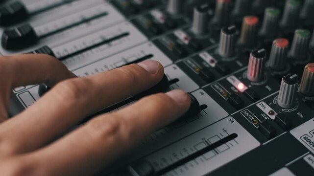 Close-up of a sound engineer's hand adjusting multiple faders on a professional audio mixing console. The shot highlights decibel markings and illuminated buttons in a recording studio.