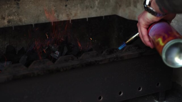 A close-up cinematic shot of a hand using a portable gas blowtorch with a blue flame to light black charcoal pieces in a metal grill barbecue, creating sparks and small flames
