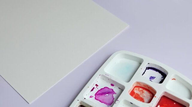 Watercolor painting workspace setup with blank paper, palette and paints appearing one by one. Art supplies arranged for creative process, artist preparing materials before painting at home studio.