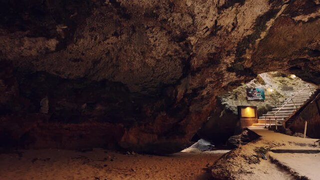 Wide angle view of a dark limestone cave interior featuring sandy floors and a lit staircase leading to the surface. Exploring hidden coastal caverns and underground tourist destinations.