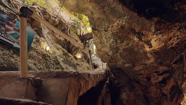Walking path inside a cavernous tunnel featuring concrete steps and metal handrails. A sign points the way to a local shop or warung within the cave system.