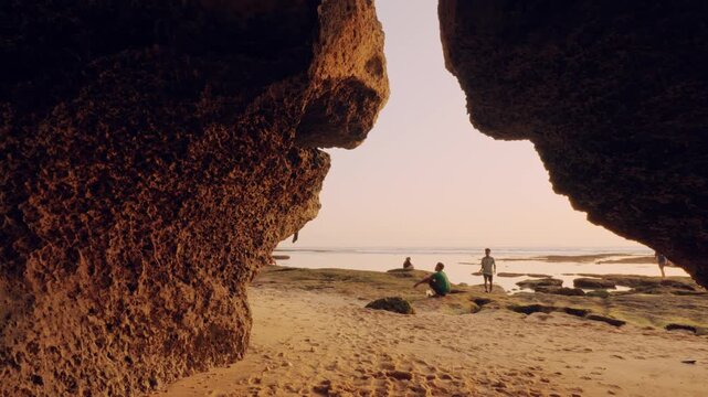 An interior perspective of a sea cave opening onto a reef beach during a warm sunset. Silhouetted rock formations create a dramatic border around the peaceful seascape.