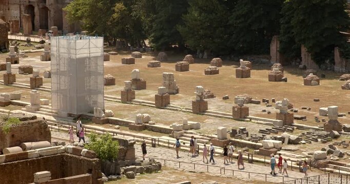 An elevated view of the ancient Roman Forum ruins in Rome, Italy, showcasing rows of historic column bases, a structure covered in white scaffolding, walking tourists, and many beautiful green trees.