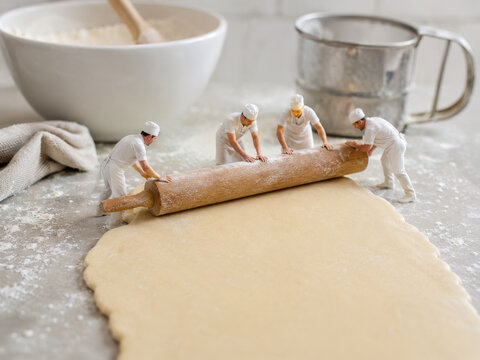 Cuatro figuras peque&ntilde;as usan un rodillo para extender masa en una cocina. Hay ingredientes y utensilios en la mesa
