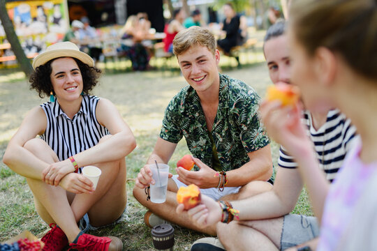 Group of young friends at summer festival, sitting on the ground and eating.