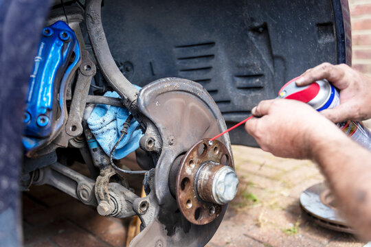 Mechanic spraying lubricant on car brake hub.Close view of a car wheel hub and brake disc during repair, with a mechanic hand holding a blue cloth and a power drill nearby in an outdoor workshop.
