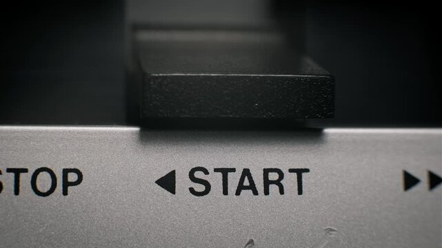 Extreme close-up of a male finger pressing the black start button on a silver vintage audio player. The Start and Stop labels are clearly visible as the mechanism begins.