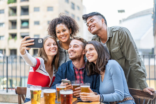 Smiling multiethnic friends taking a cheerful beer selfie together at an outdoor city cafe terrace. Young adults gather close during happy hour. Social media concept